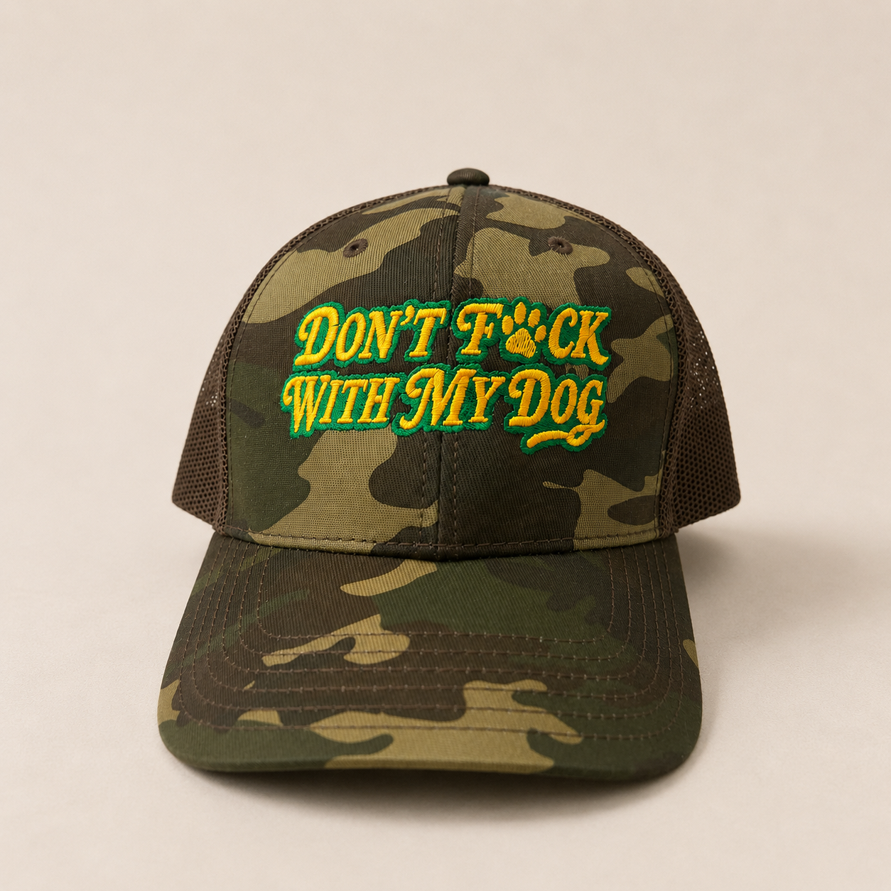 A camouflage trucker hat with mesh back panels and embroidered text reading 'DON'T F*CK WITH MY DOG' in yellow and green, shown on a plain light beige background.